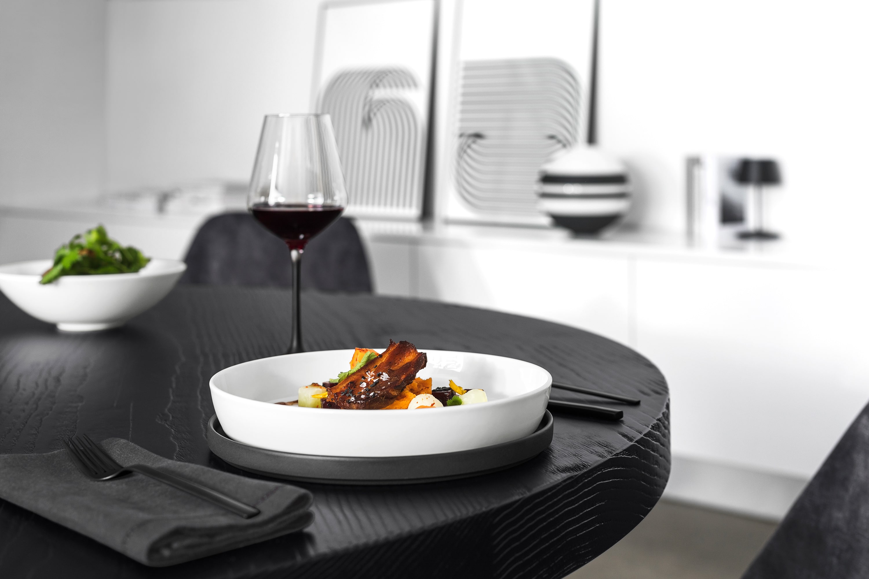 White La Boule as fine dinner A white La Boule plate with food and a glass of red wine on a dark wooden table with modern decor in the background.