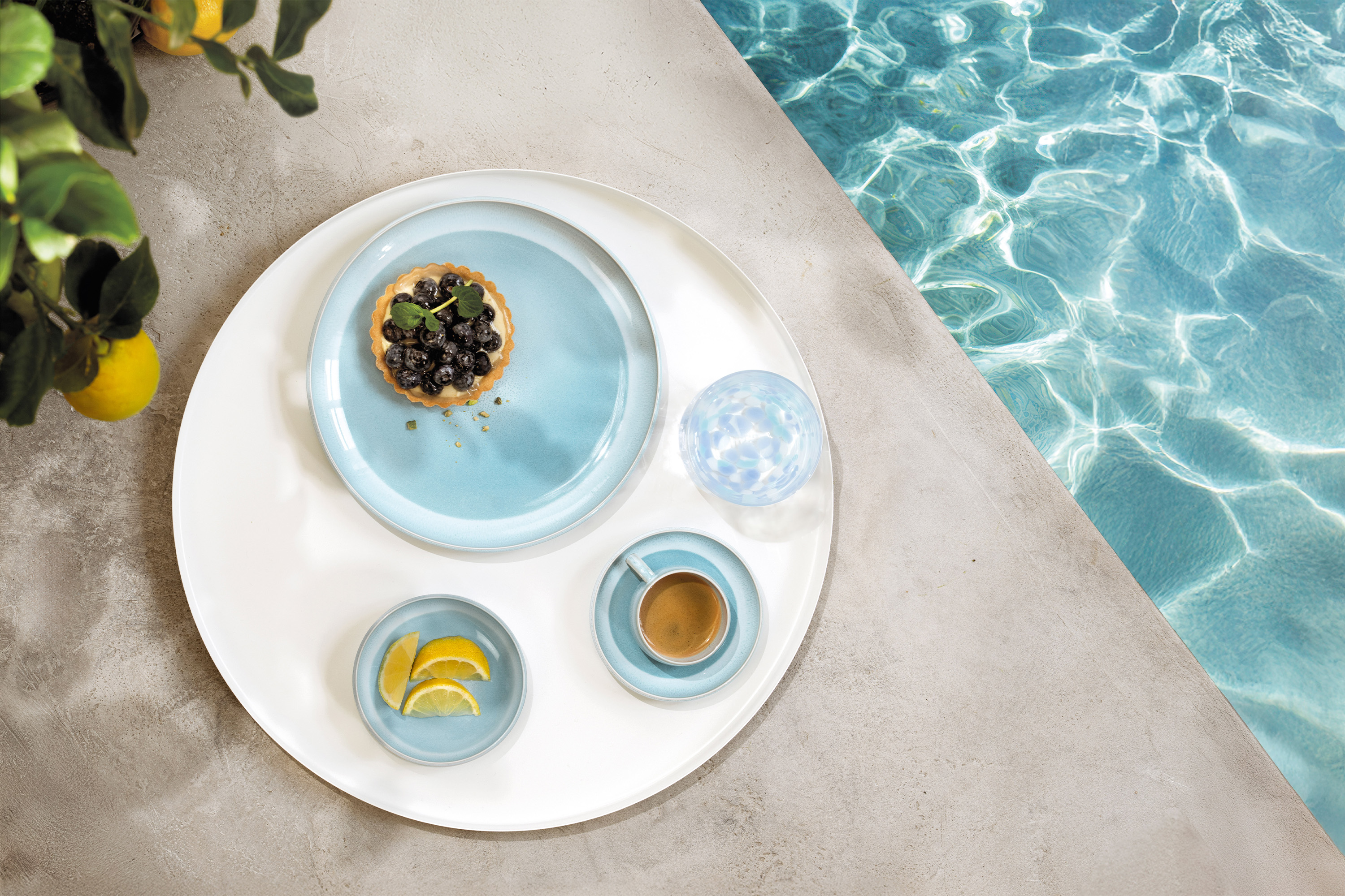A round tray with tableware from the Perlemor Aqua collection A round tray with tableware from the Perlemor Aqua collection and a glass of water, at the edge of a pool.