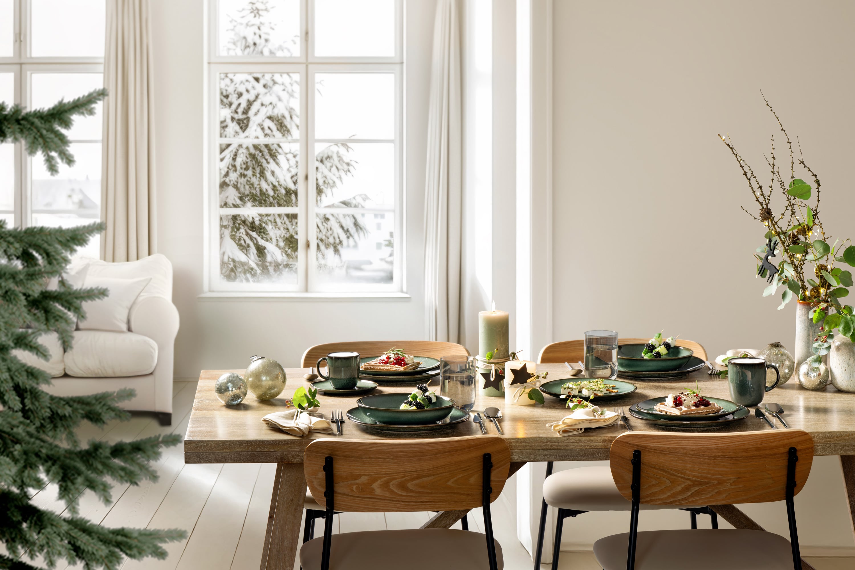 Une table à manger en bois pour quatre personnes avec des assiettes, des tasses et des bougies Lave Vert dans une pièce lumineuse donnant sur la neige à l'extérieur.