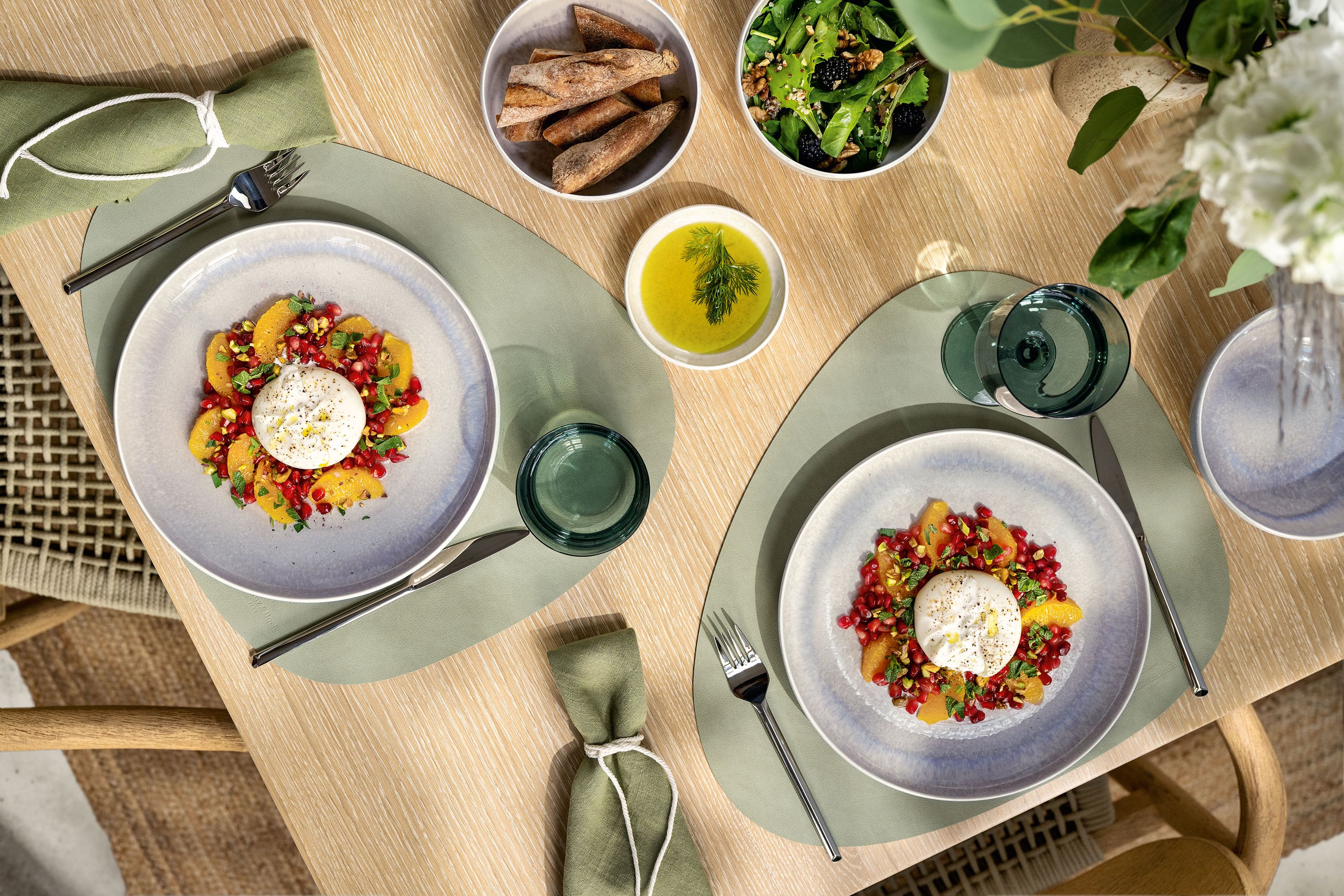 Zwei Perlemor Teller in beige mit Salat und Burrata-Käse Zwei Teller Salat mit Burrata-Käse, serviert mit Brot, grünem Salat und einer Schüssel Olivenöl auf einem Esstisch.