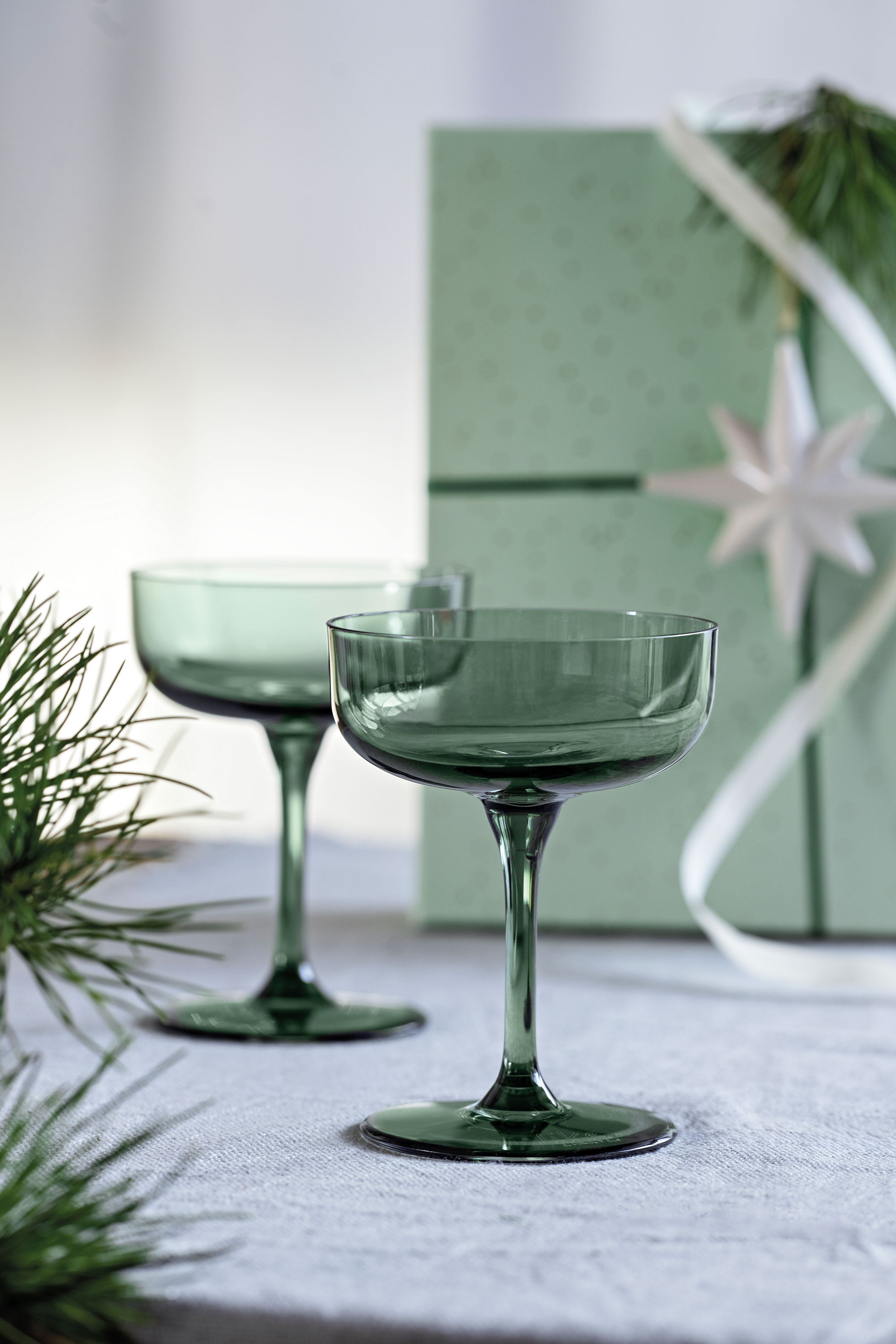 Like Sage glass ideal as a gift Two green cocktail glasses on a white surface with a green gift box and fir branches in the background.