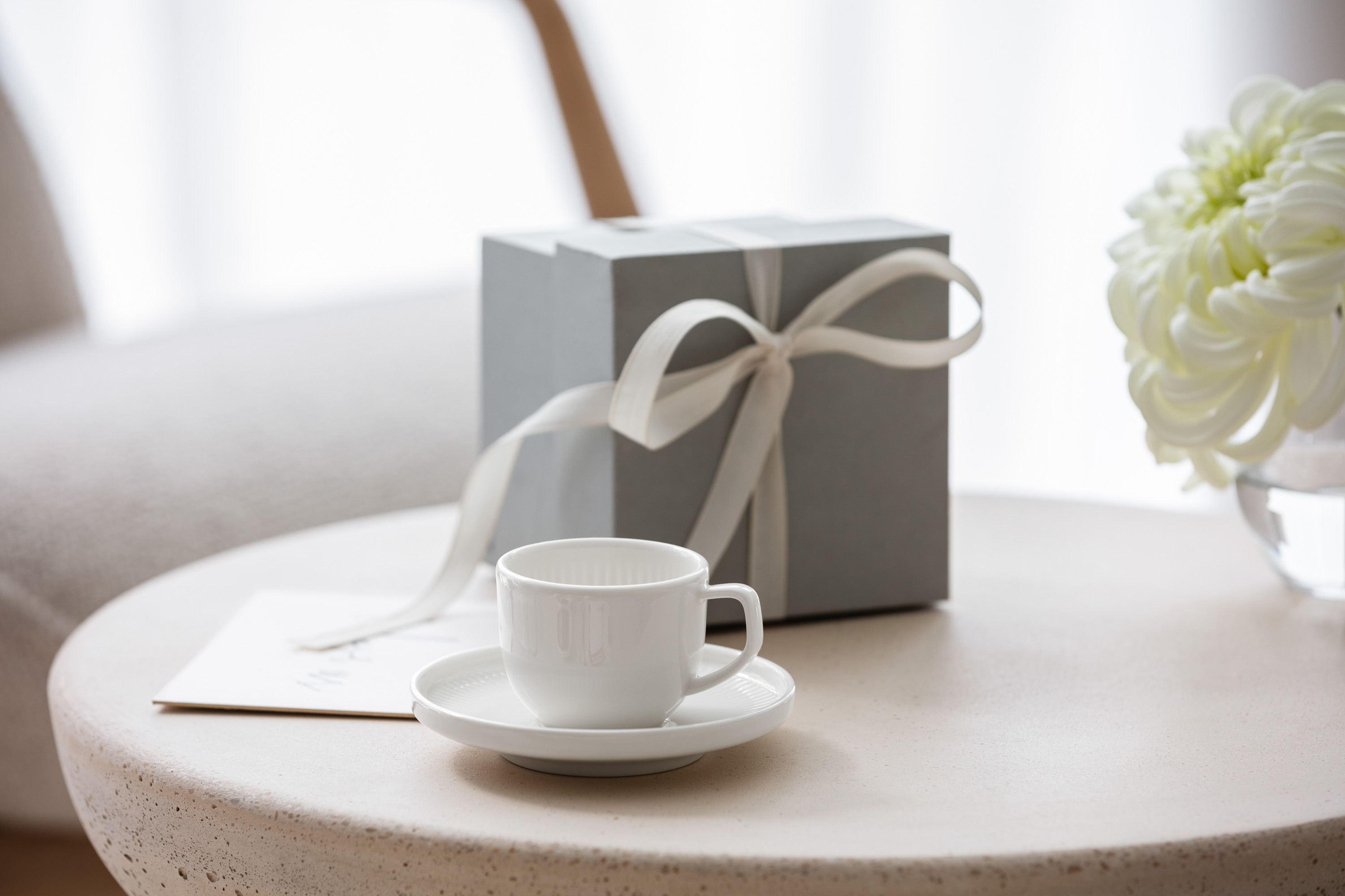 Afina espresso cup and saucer with a gift box A white Afina cup on a saucer, a gift box with a bow and a bouquet of white flowers on a round table.