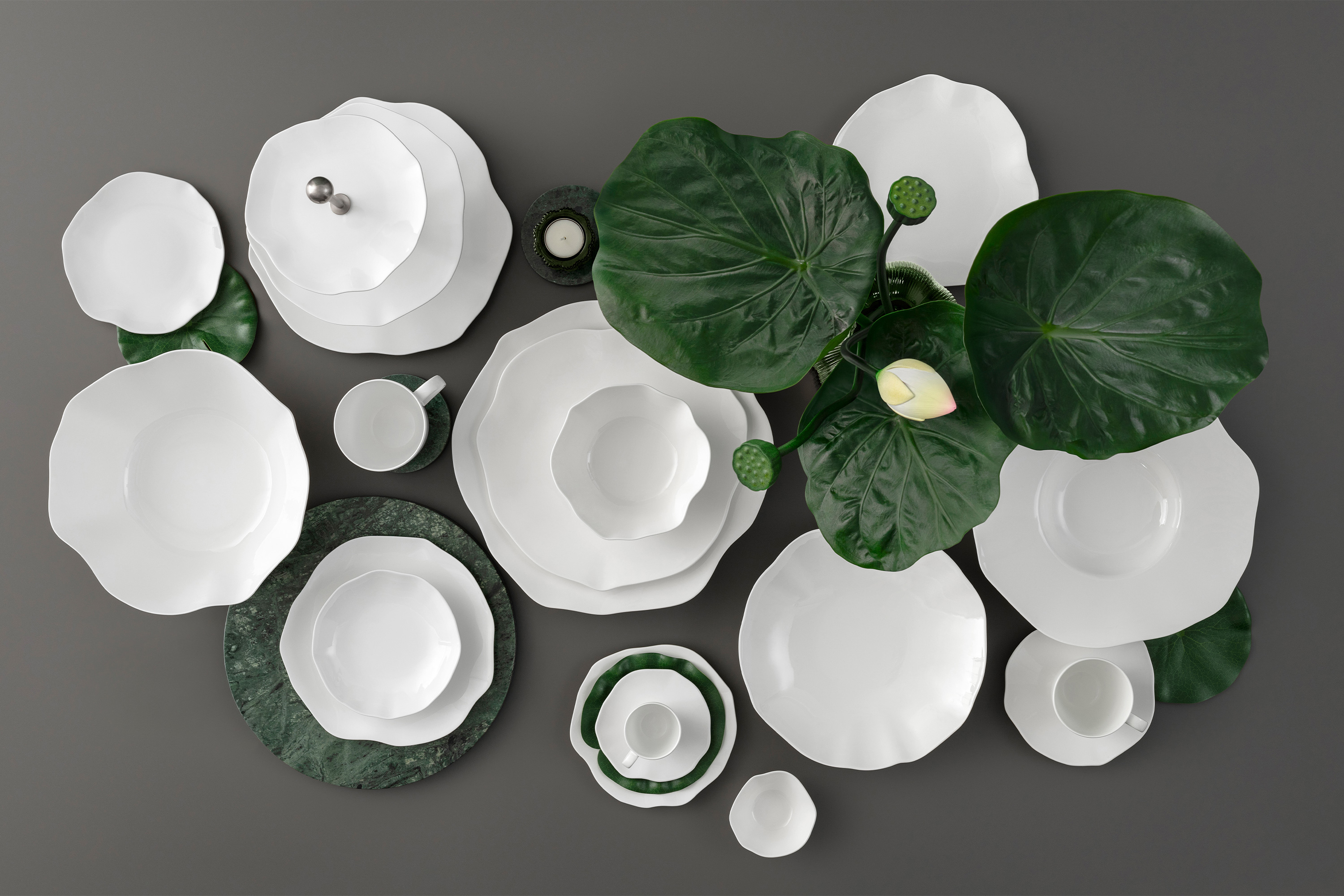 Weiße Geschirrteile der Kollektion Pura auf einer dunklen Fläche mit großen grünen Blättern und einer einzelnen weißen Blume als Dekoration.