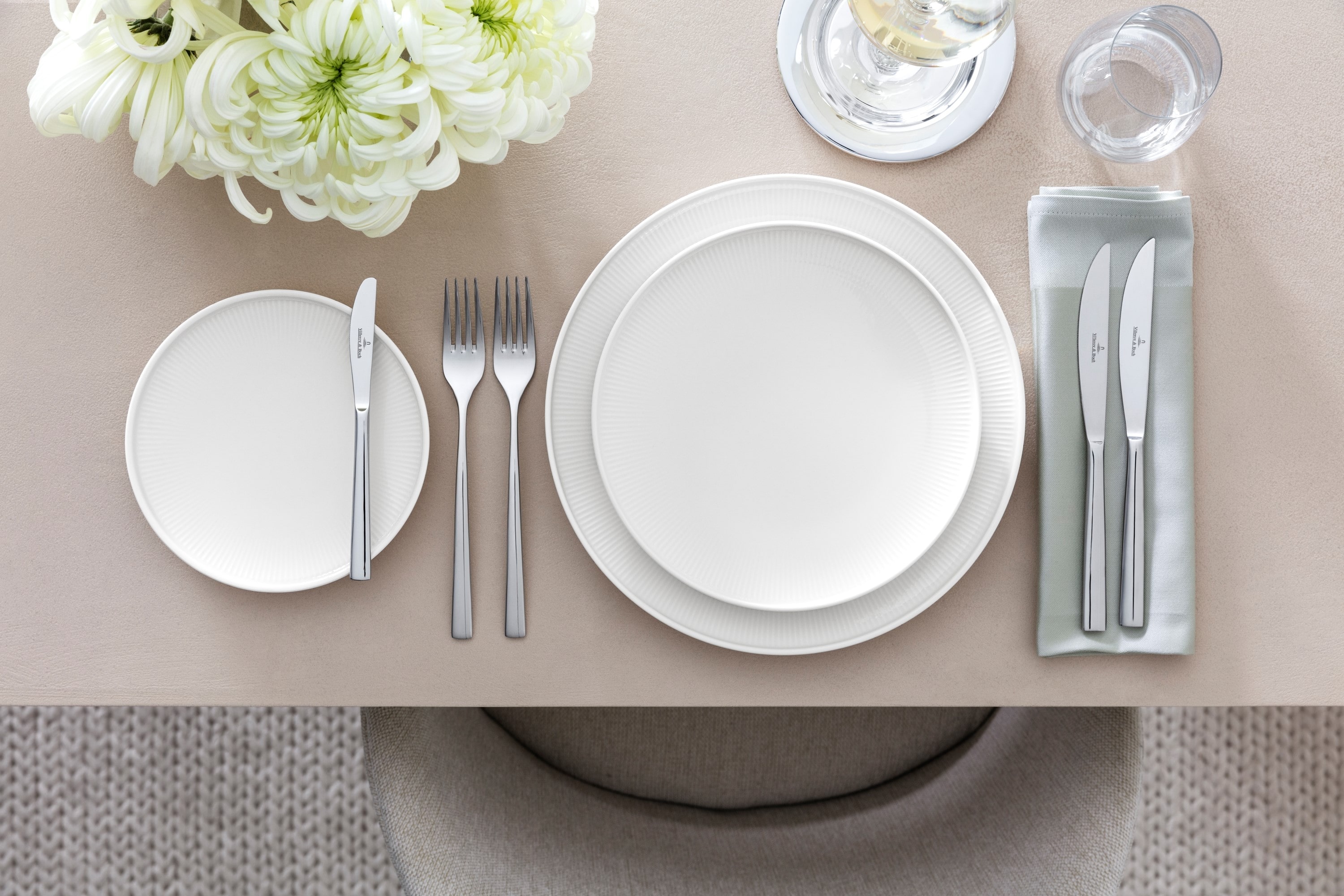 Table setting with Afina dinnerware and Piedmont flatware Sophisticated table arrangement with the Piemont collection from Villeroy & Boch, including a white dinner plate, a salad plate, a flatware set, glassware, and a floral centerpiece on a beige tablecloth.