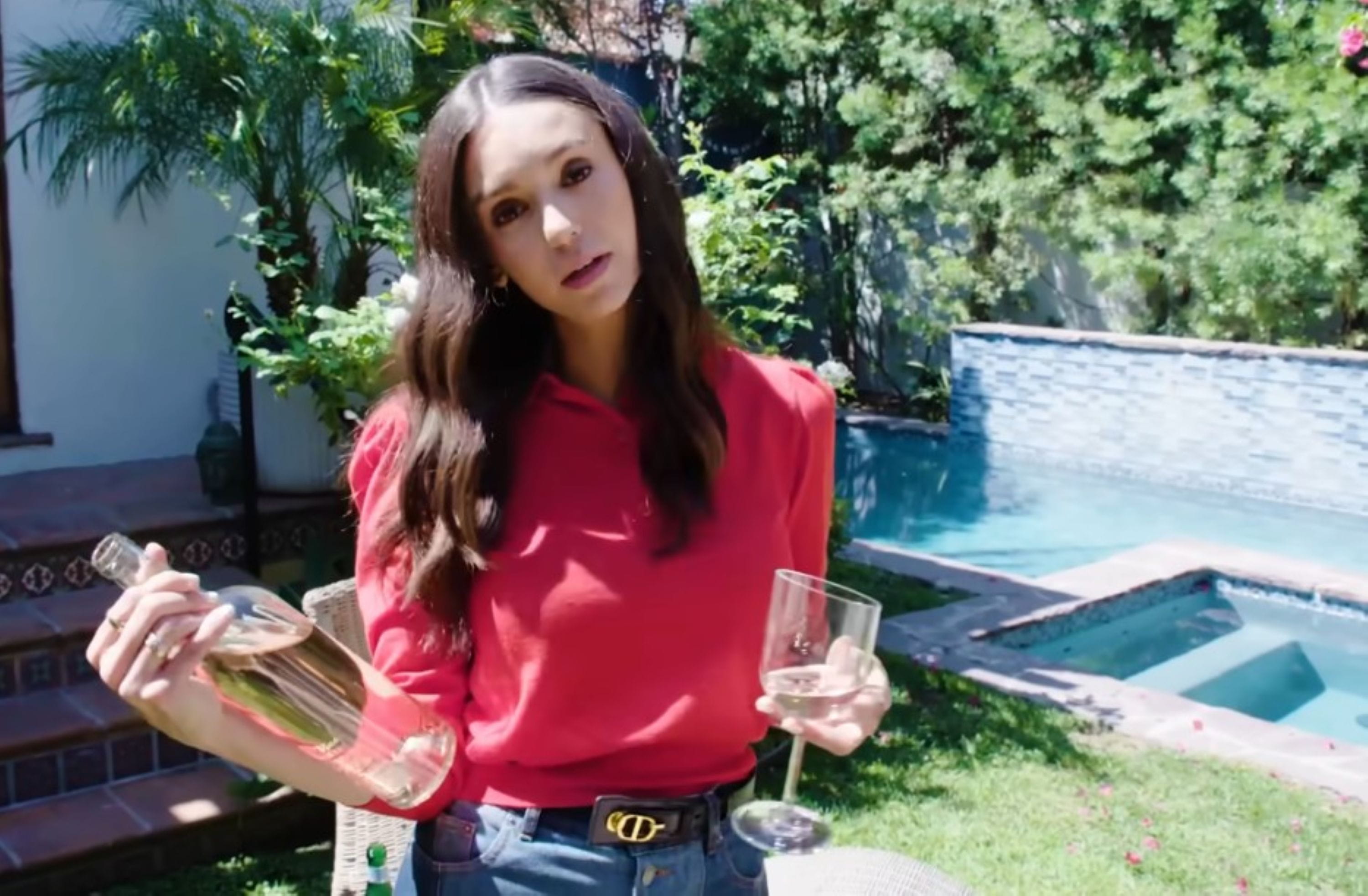architecture-digest-as-seen-on Woman in a red top stands in a garden holding a wine bottle and glass, with wavy hair. A pool, greenery, and a patio create a relaxed outdoor setting.