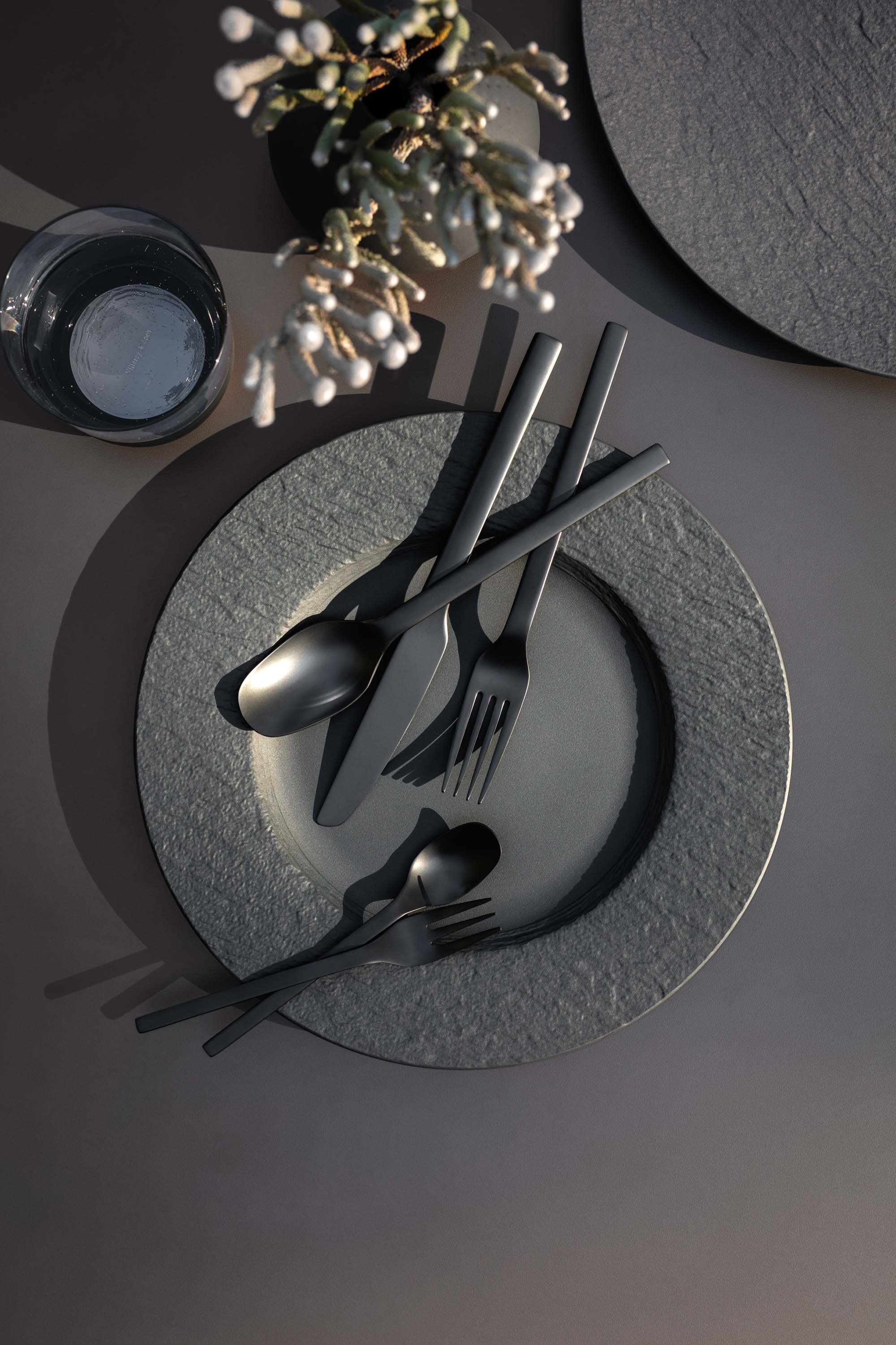Des couverts de table Manufacture avec des assiettes plates Manufacture Une décoration de table élégante avec des couverts de table Manufacture modernes sur des assiettes Manufacture noires avec une branche décorative et un verre d’eau.