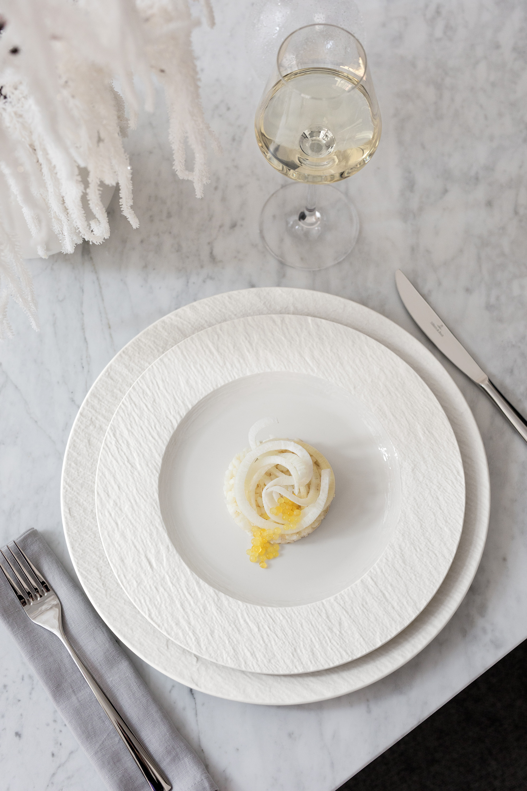 Un plato blanco Manufacture con un pequeño postre y una rodaja de limón, al lado de un cuchillo, un tenedor y una copa de vino blanco sobre una mesa gris.