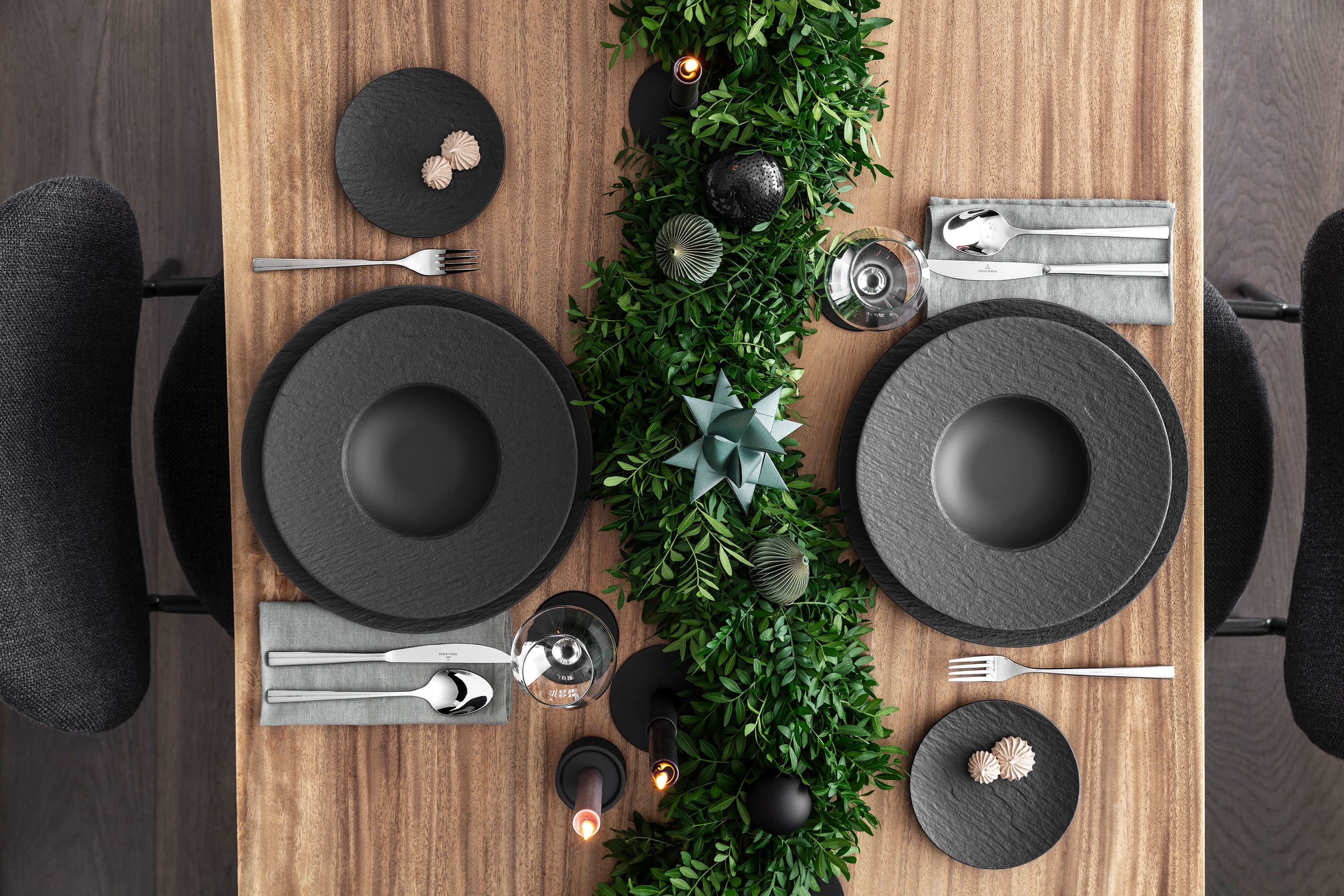 Une élégante table de fête avec des assiettes noires Manufacture, des couverts de table et des décorations de fête vert foncé sur une table en bois.