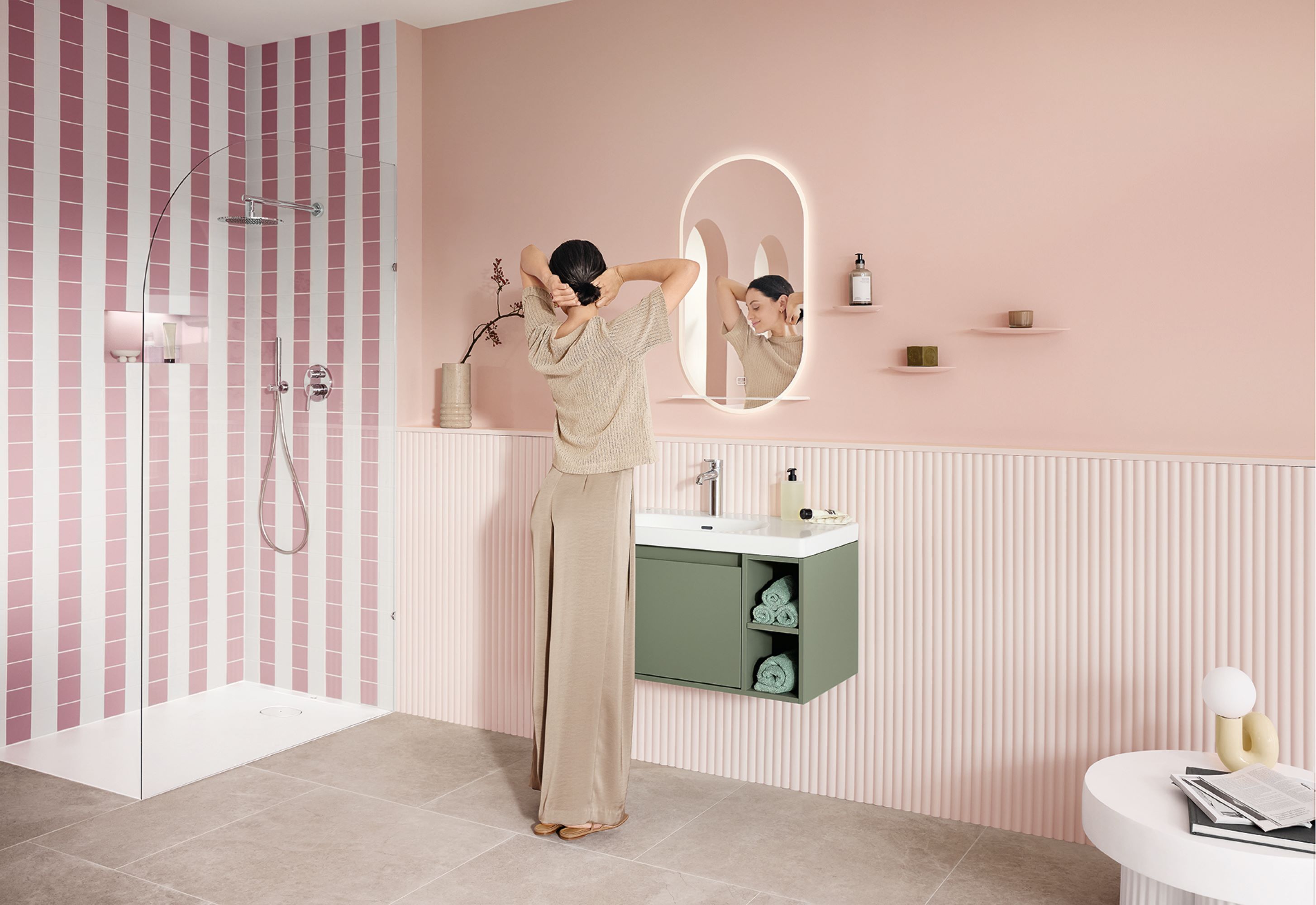Praktyczna umywalka z kolekcji Skyla i More To See Lite+ firmy Villeroy & Boch. Kobieta wyciąga się przed lustrem przy umywalce w stylowo urządzonej pastelowo-różowej łazience. Umywalka składa się z białej szafki umywalkowej z jasnozieloną szafką podumywalkową i chromowanej armatury z kolekcji Skyla. Podświetlane, owalne lustro More To See Lite+ ma praktyczną półkę w kolorze białym.