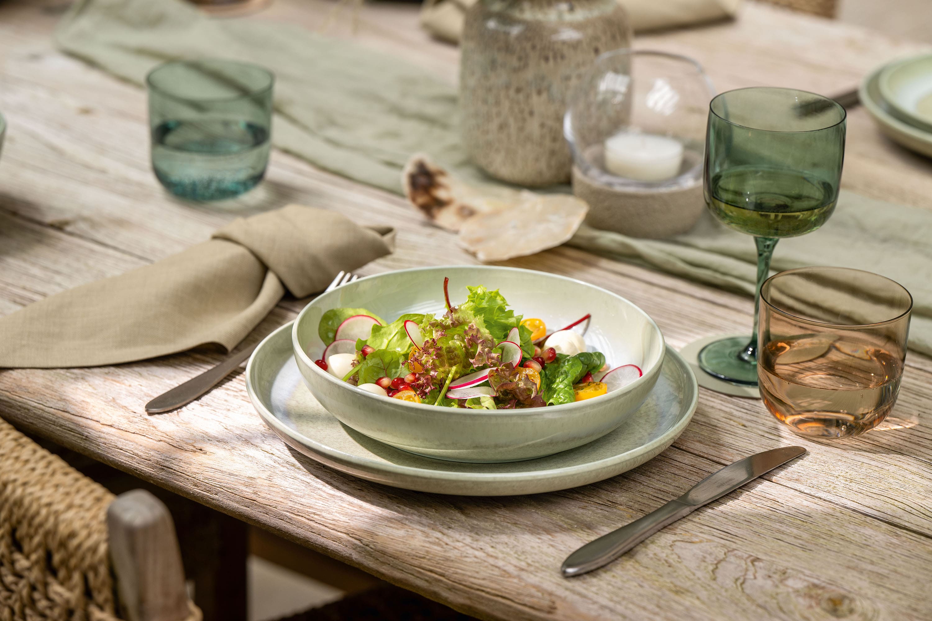 A bowl of salad sits on a set dining table with Perlemor glasses, plates, and beige napkins.