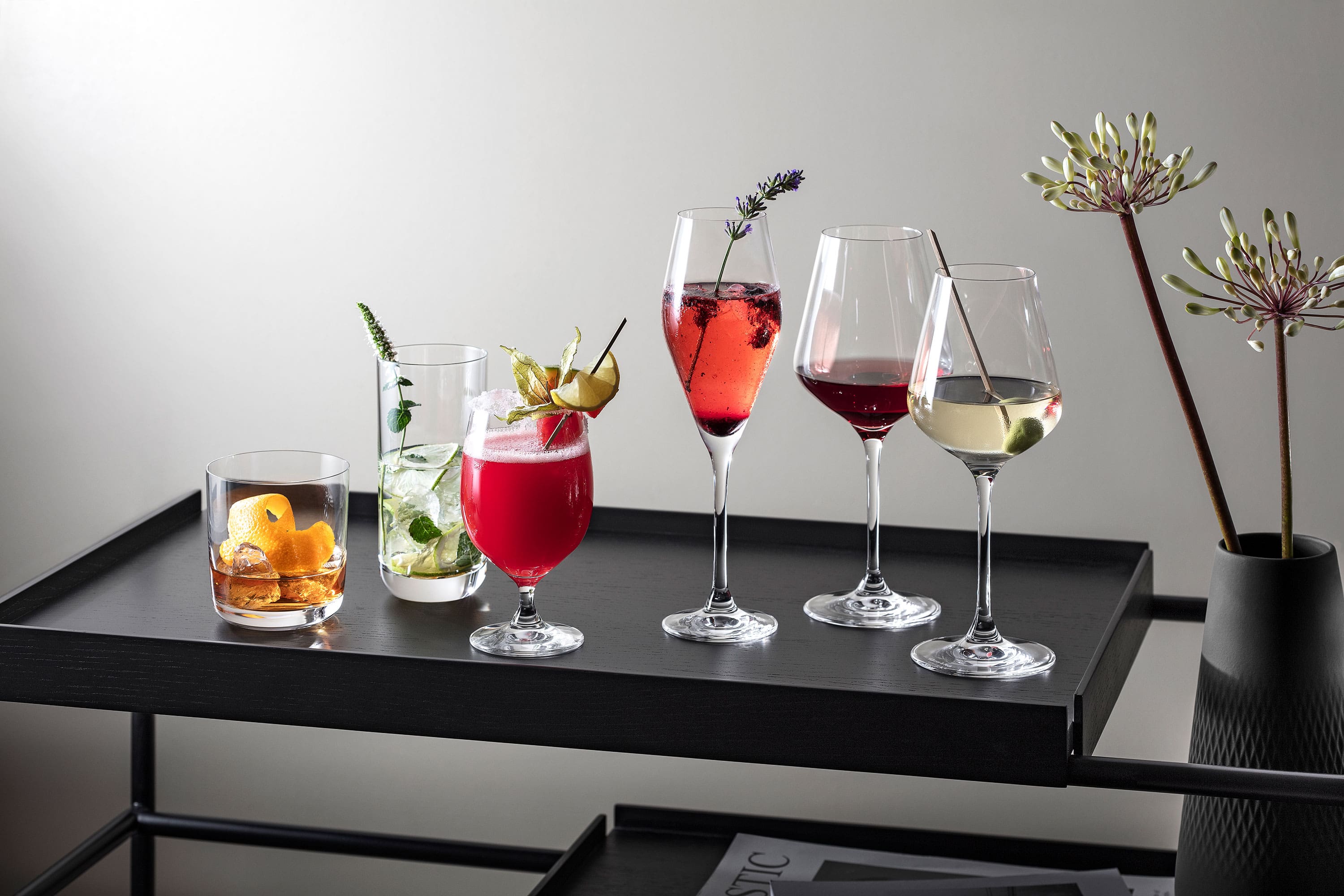 Various drinks in La Divina glasses, presented on a modern black tray, garnished with herbs and fruit.