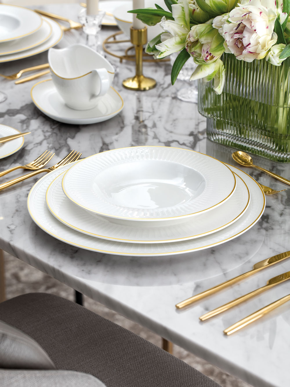 Un service de table formel avec des assiettes blanches, des couverts en or, une saucière blanche et une composition florale sur une table en marbre.