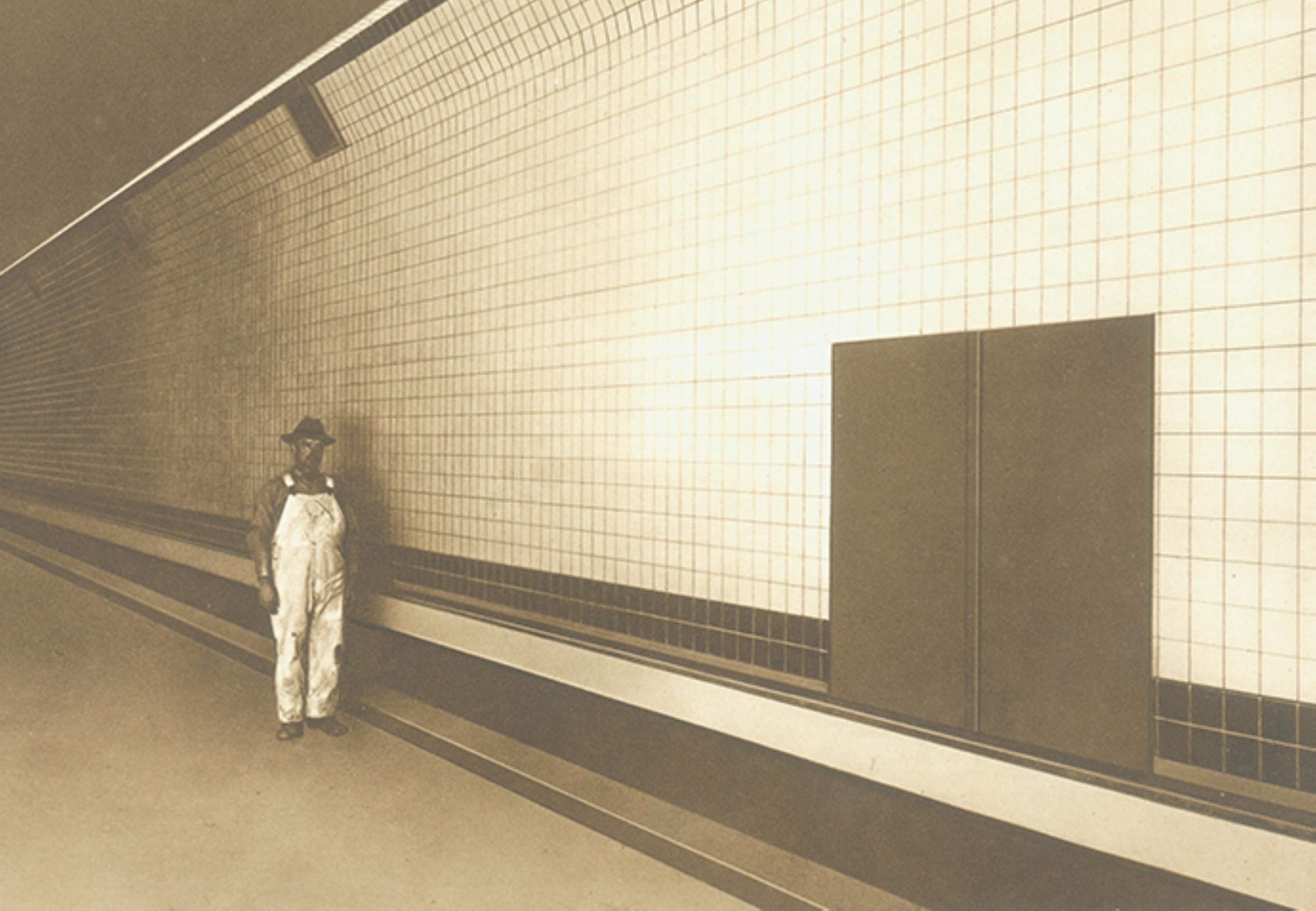 vb-holland-tunnel-tiles A person in overalls and a hat stands next to Villeroy & Boch tiles in the minimalist, empty corridor of the Holland Tunnel.