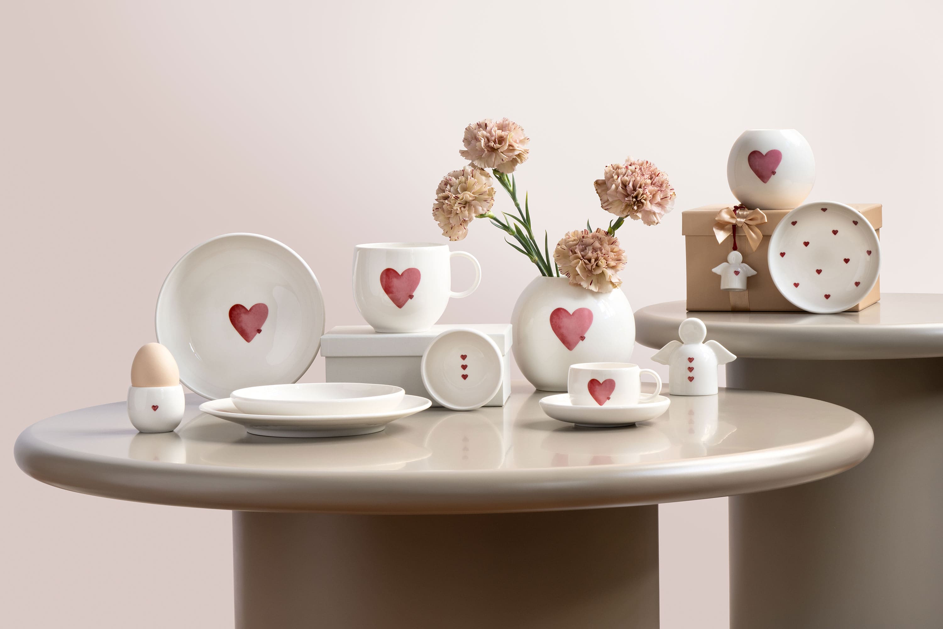 Vajilla de cerámica de With Love con motivos de corazones rojos, dispuesta sobre mesas redondas con flores y una caja de regalo.